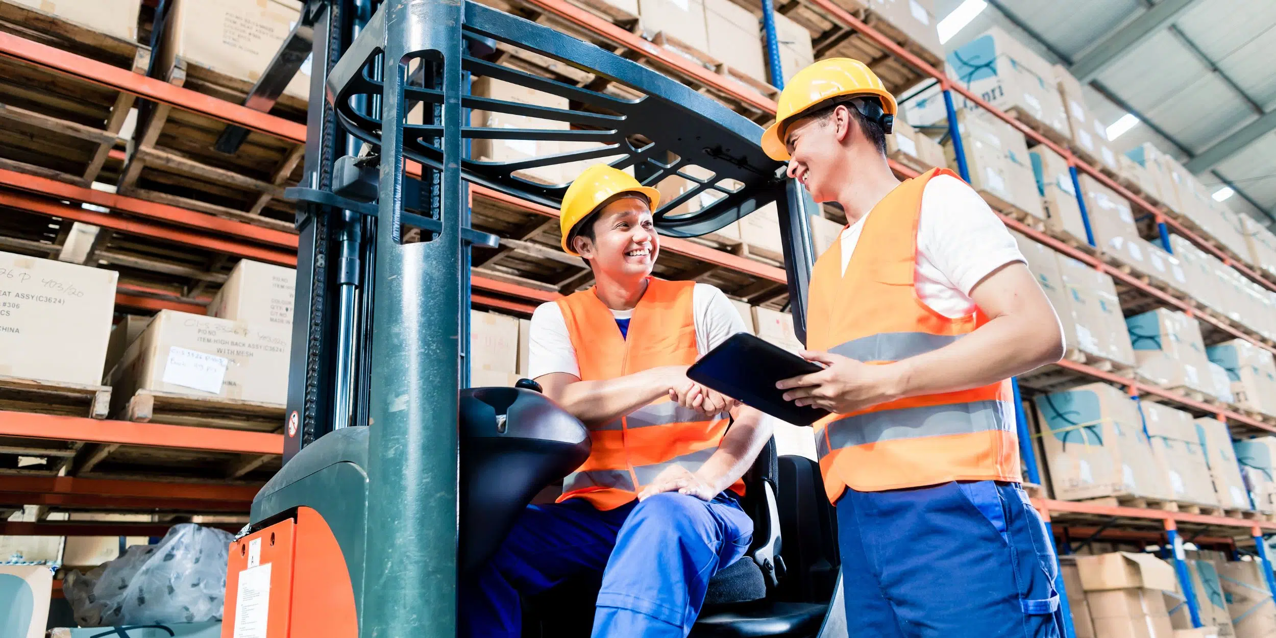 Symbolbild für das Angebot "LogiPLUS": Zwei Lagerarbeiter in orangefarbenen Sicherheitswesten und Schutzhelmen stehen in einem großen Lager zwischen hohen Regalen. Einer sitzt auf einem Gabelstapler, der andere hält ein Tablet. Beide lächeln und geben sich die Hand, als würden sie sich über erfolgreiche Arbeit austauschen.